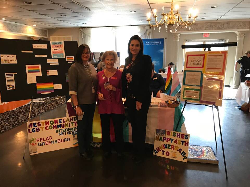 Three women are standing in front of a table for PFLAG and the Westmoreland LGBTQ Interfaith Community.